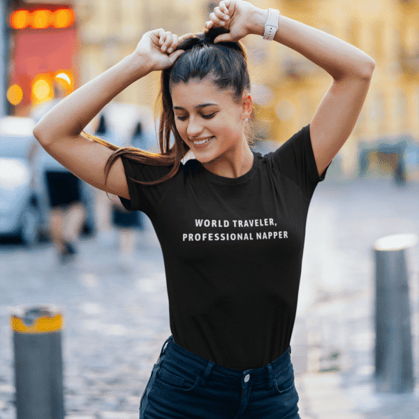 Woman smiling in the street wearing a black Travel T-Shirt with the text “World Traveler, Professional Napper.”