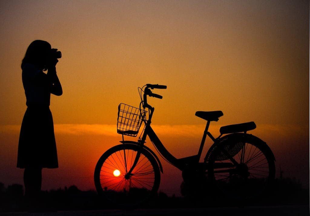 A silhouette of a woman photographer and a bicycle against a vibrant sunset background.