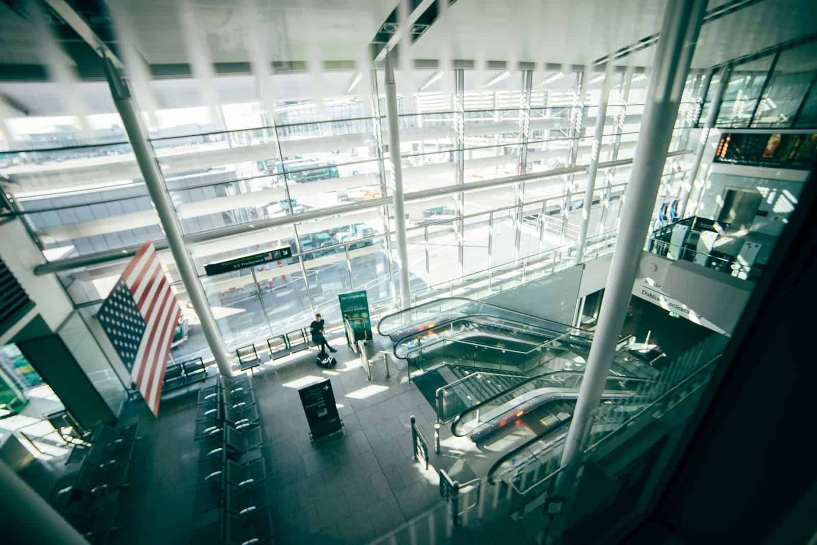 A bright, spacious airport terminal featuring expansive glass walls, escalators, and American flag.